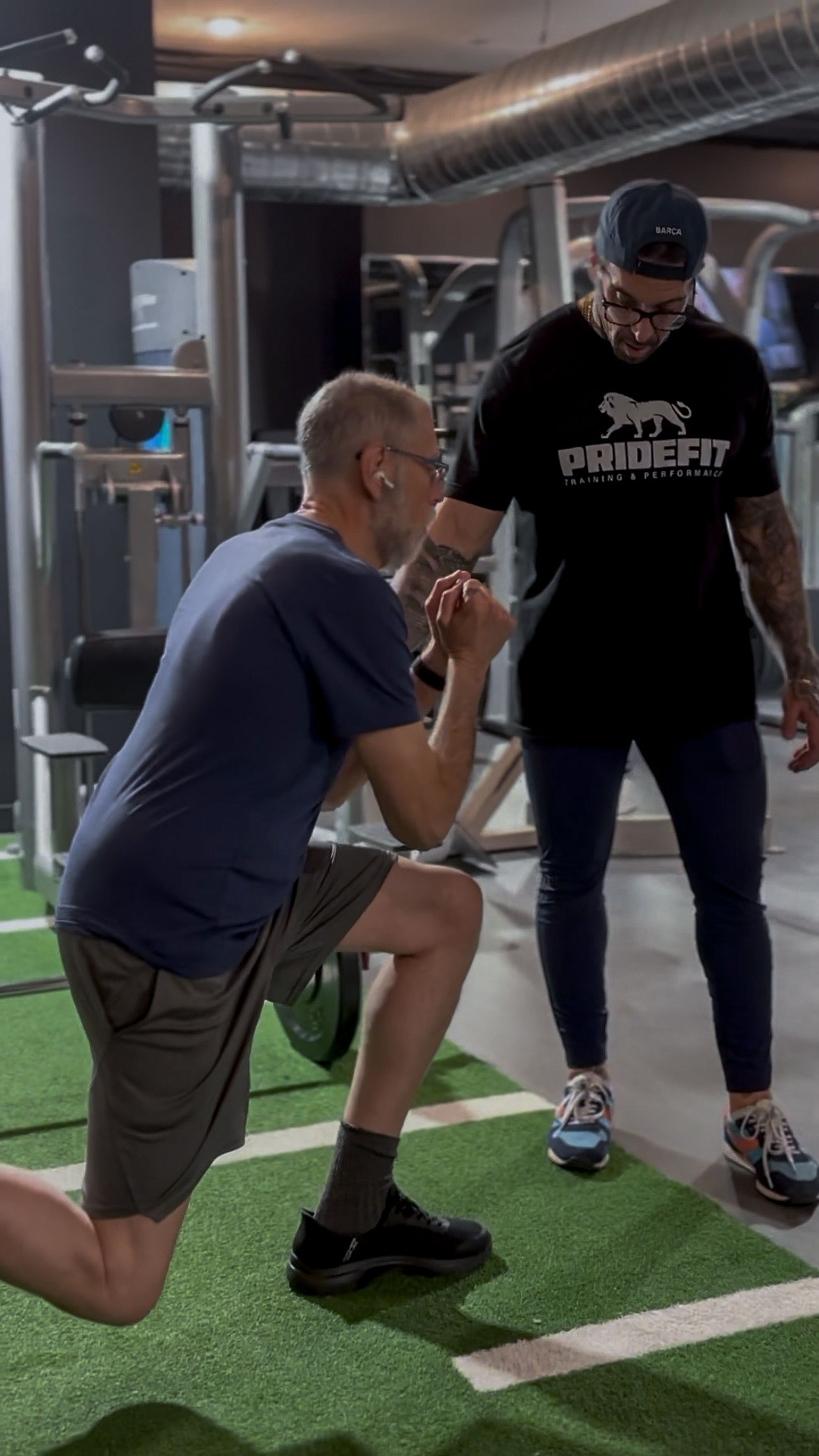 Joe Ferrero, in a PrideFit shirt and backwards cap, coaching a member through a kneeling split-squat on the turf